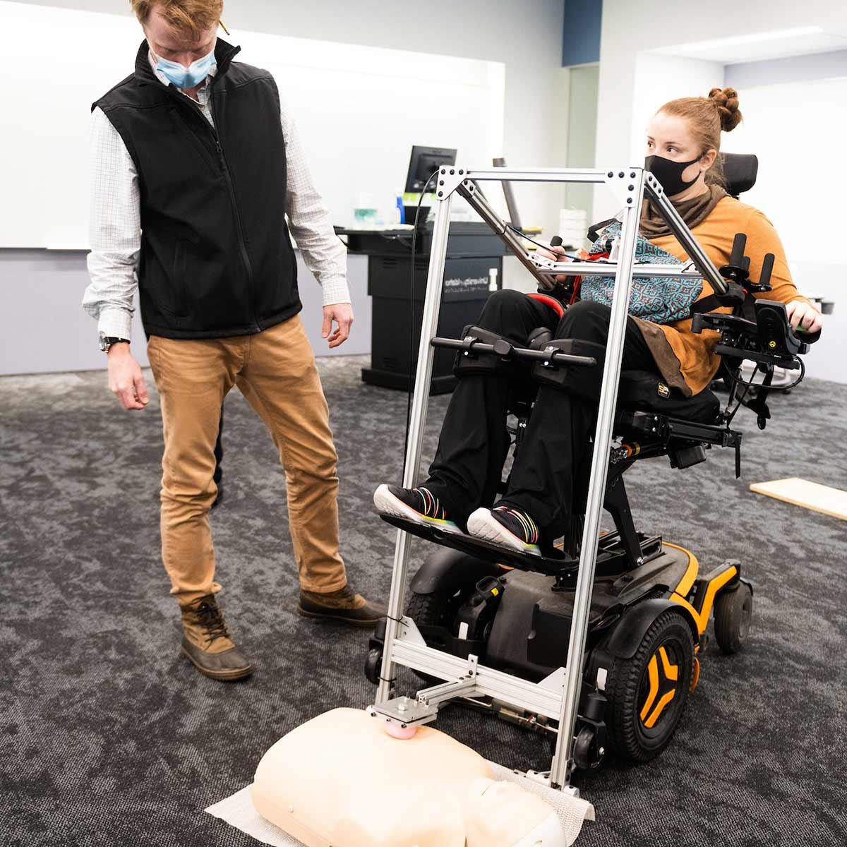 工科学生帮助坐在轮椅上的学生使用辅助心肺复苏装置, 哪一个使用杠杆动作来增加力量和实现必要的胸部压迫.